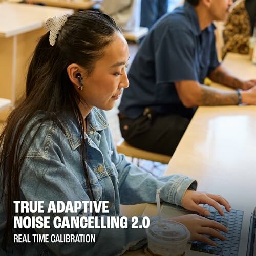Woman in denim jacket using laptop at cafe, wearing earbuds.